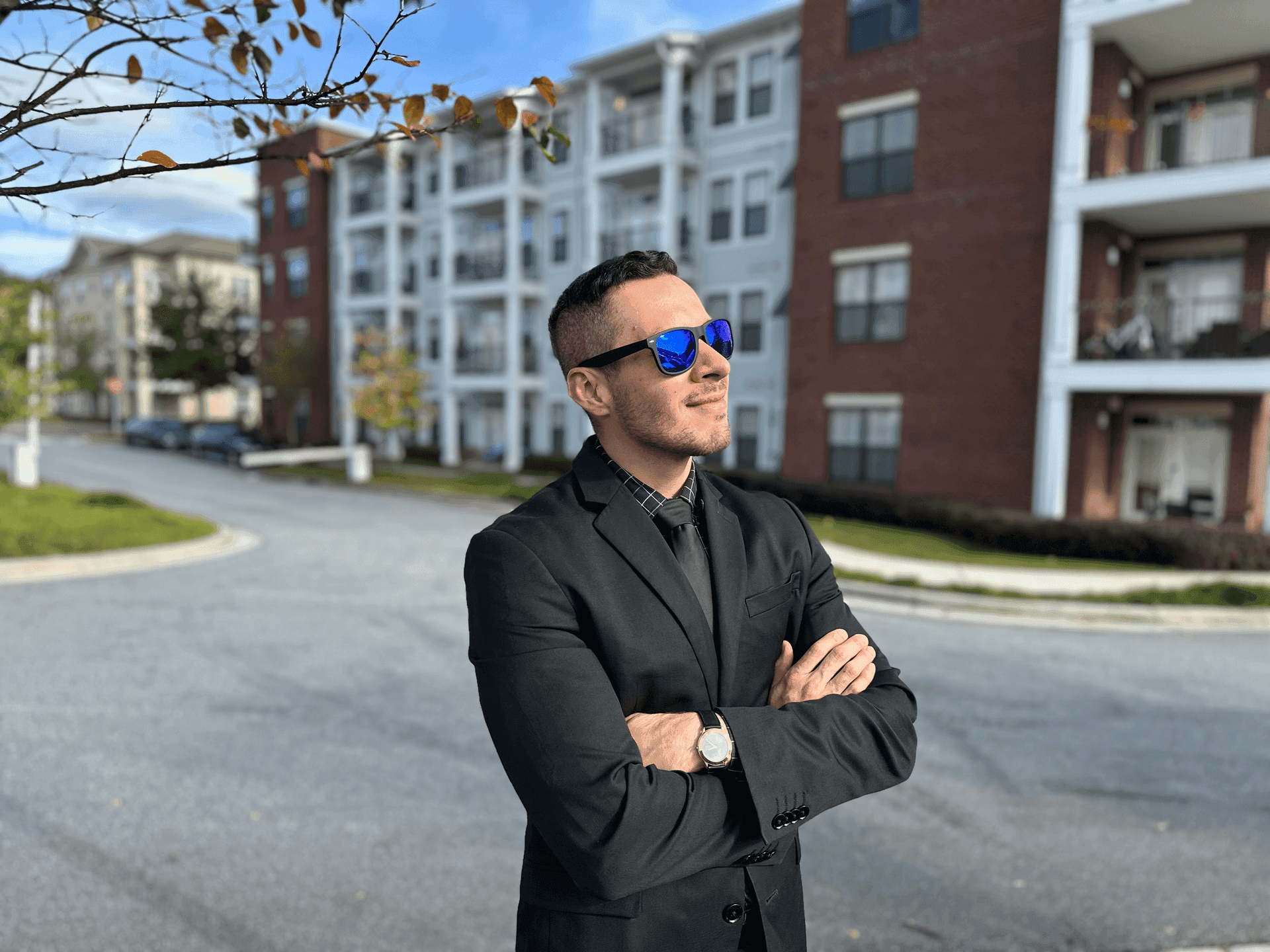 Confident person in a black suit and blue sunglasses standing outside apartment buildings on a sunny day.