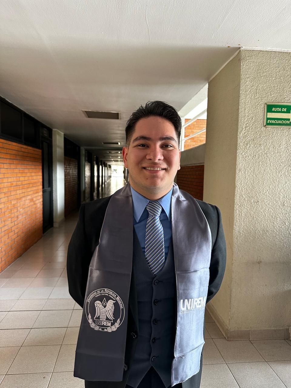 Person in a suit with a graduation sash standing in a hallway with brick and beige walls.