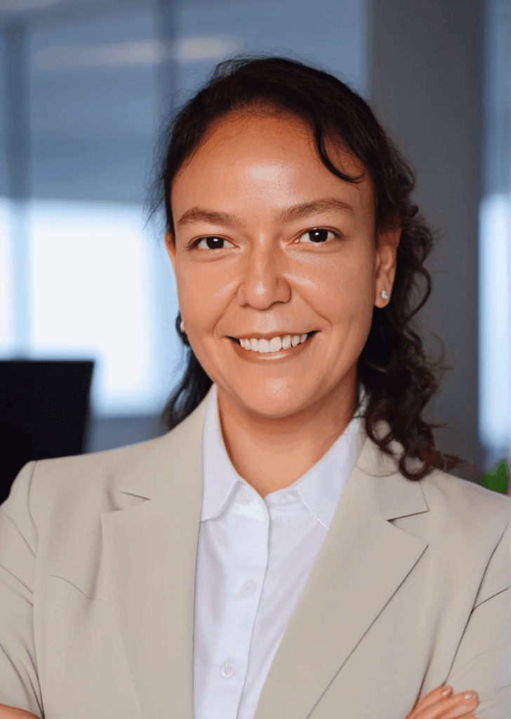 Smiling person in a beige blazer with arms crossed in a modern office setting.